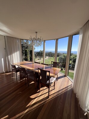 A dining area with a large wooden table and chairs, large windows with curtains, and a chandelier. - The Cutting Estate Mount Panorama (Mount Panorama)