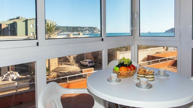 Un balcón con una mesa blanca dispuesta con fruta, tazas de té y un pastel, con vistas a una zona costera con edificios y cielo despejado.