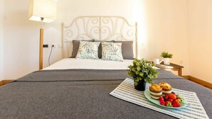 A neatly made bed with a grey blanket, a white headboard, and two pillows with leaf patterns. A small table with a plant and a plate of strawberries and muffins is visible.