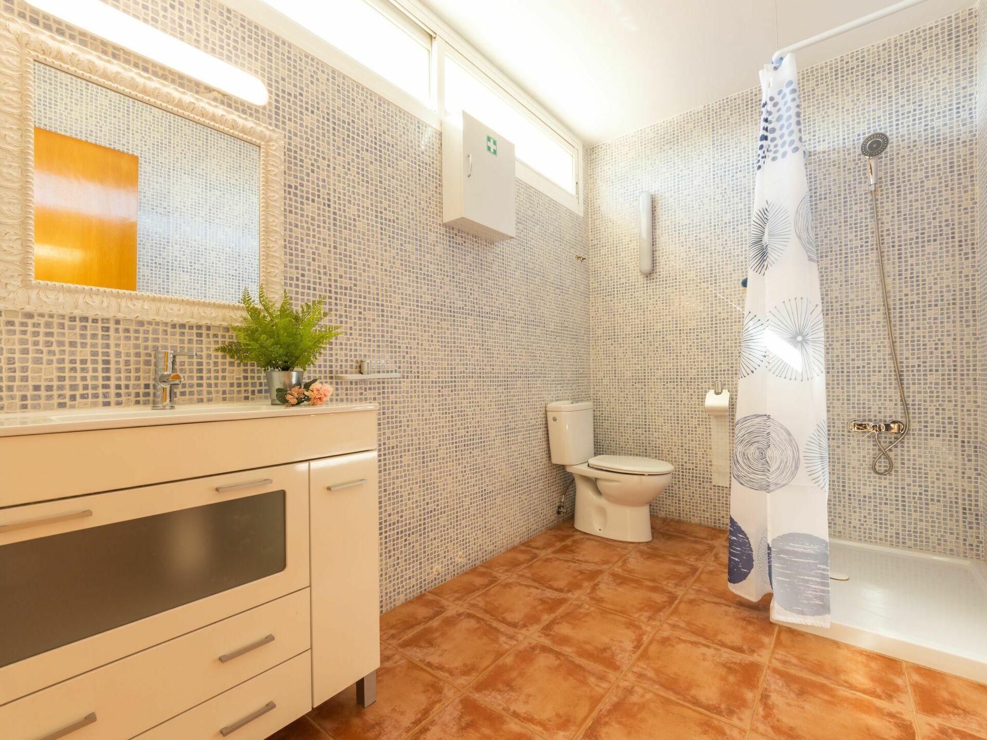 A bathroom with a tiled floor, a toilet, a shower with a curtain, a sink with a mirror, and a plant on the counter.