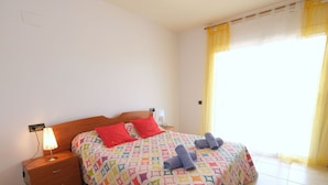 A neatly made bed with a colorful geometric-patterned bedspread, two red pillows, and rolled towels on the bed. There is a wooden nightstand with a lamp, a small drawer, and a chair with a plaid cushion. The room has white walls, a window with yellow curtains, and tiled flooring.