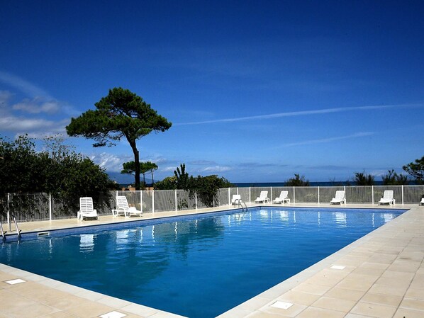A rectangular swimming pool with a tiled deck, surrounded by white lounge chairs and a white fence.
