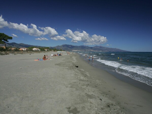 A beach with people sunbathing and swimming, a clear sky, and mountains in the background.