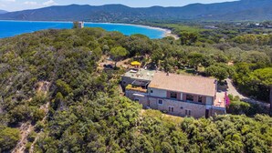 Una villa con tejado de baldosas, rodeada de abundante vegetación, con vistas a una zona costera y una perspectiva despejada del mar y montañas distantes.