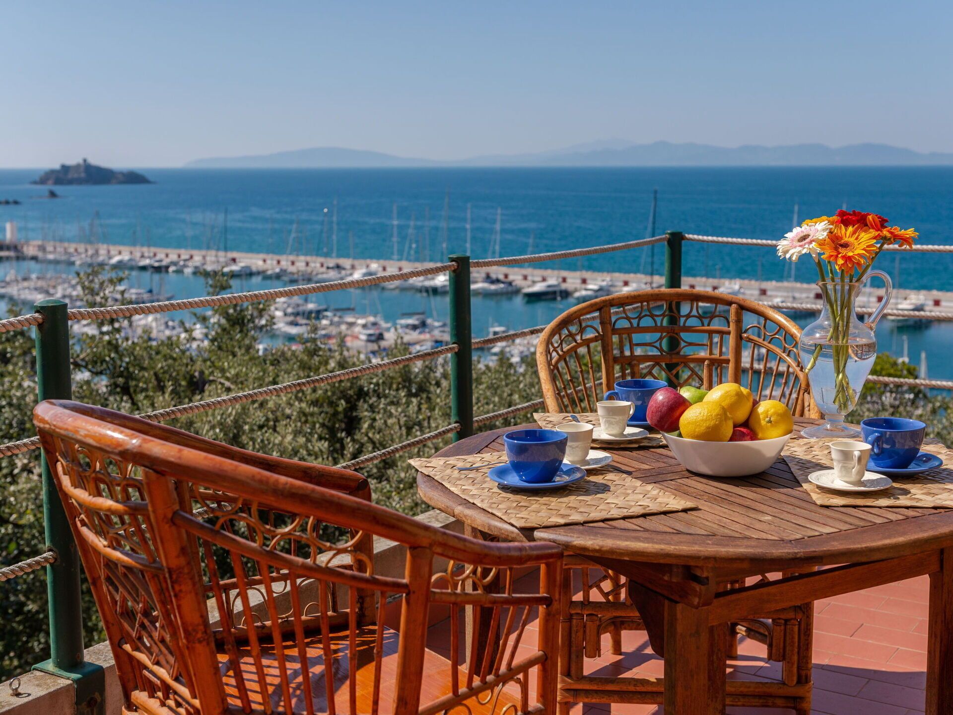 A wooden table with a bowl of fruit, a vase of flowers, and blue cups on a balcony overlooking a marina with boats and distant mountains.