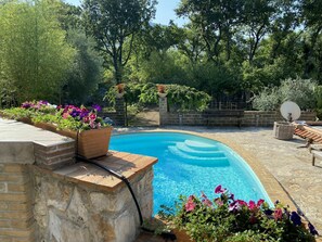 A swimming pool surrounded by a stone deck, with a brick wall on one side and a wooden deck on the other. There are flower beds with colorful flowers along the edge of the pool. - Stunning private villa for 8 guests with WIFI, private pool, TV, patio and pets allowed (Itri)