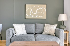 A grey sofa with white cushions, a wooden coffee table, a framed abstract artwork, and a lamp on a wooden side table.