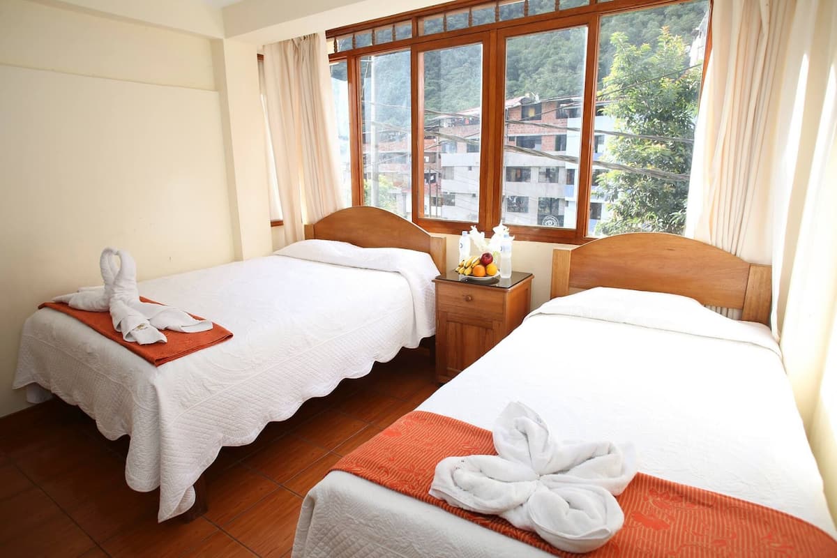 A hotel room with two beds, each with a white bedspread and a red blanket. There is a window with curtains, and a nightstand with a fruit bowl and a bottle.