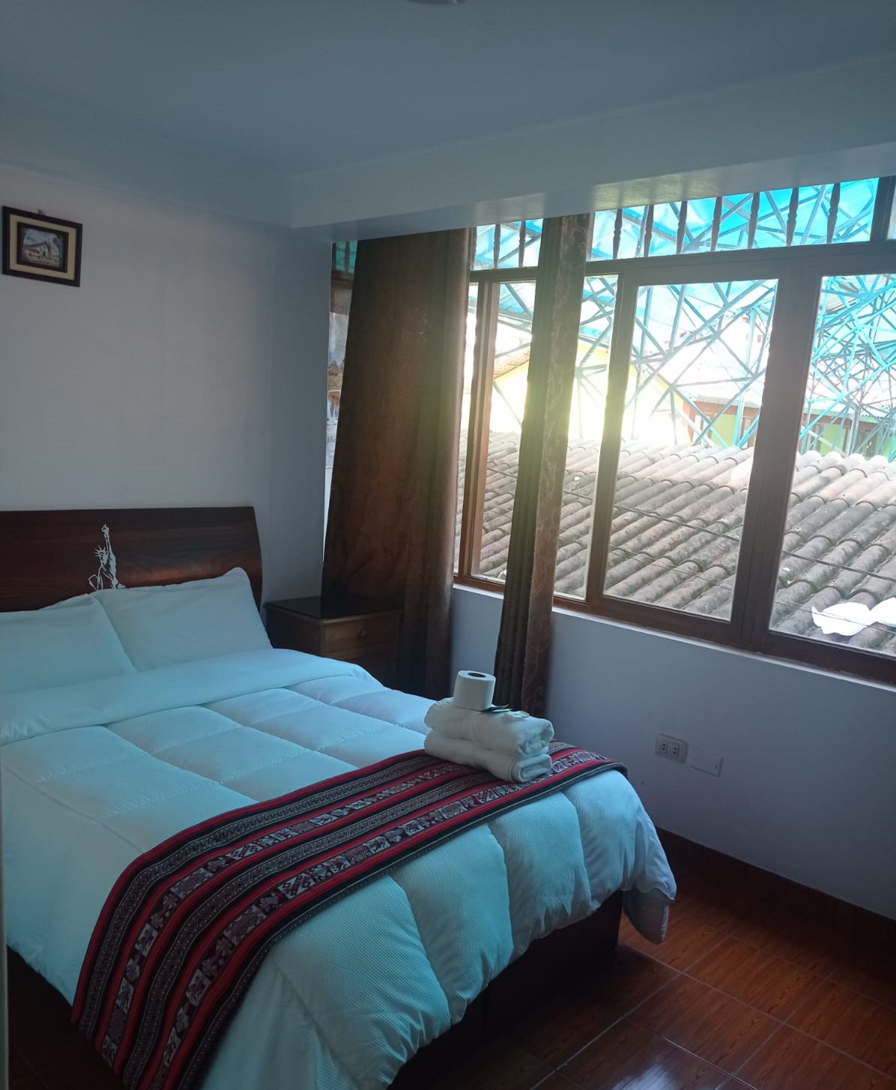 A neatly made bed with a patterned blanket, a wooden headboard, and a window with a view of a building with a blue roof.