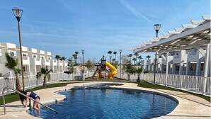 A large outdoor swimming pool with a slide, surrounded by white buildings and palm trees.