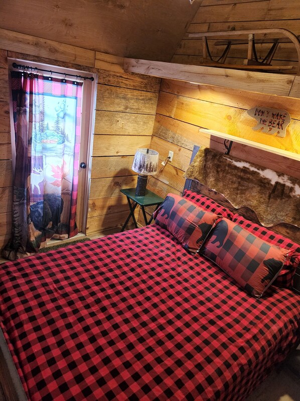 Une chambre chaleureuse dans un chalet, avec une couverture à carreaux sur le lit, une lampe sur une petite table et une pancarte sur le mur indiquant : « Ne réveillez pas l’ours ».