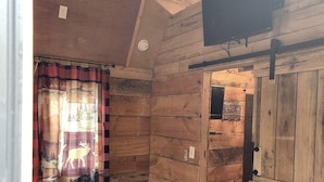 A rustic cabin room with a checkered bedspread, a ceiling fan, and a wooden door leading to a bathroom. - Lazy Bear Retreat by the Creek  (Pickens)
