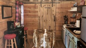 A rustic cabin interior with a wooden ceiling, a chandelier, a fur-covered chair, a kitchen area, and a dining table with stools. - Lazy Bear Retreat by the Creek  (Pickens)