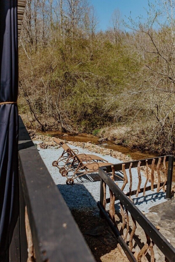 Eine Holzterrasse mit Metallgeländer, die einen natürlichen Landschaftsbereich mit Bach und vereinzelter Vegetation überblickt.