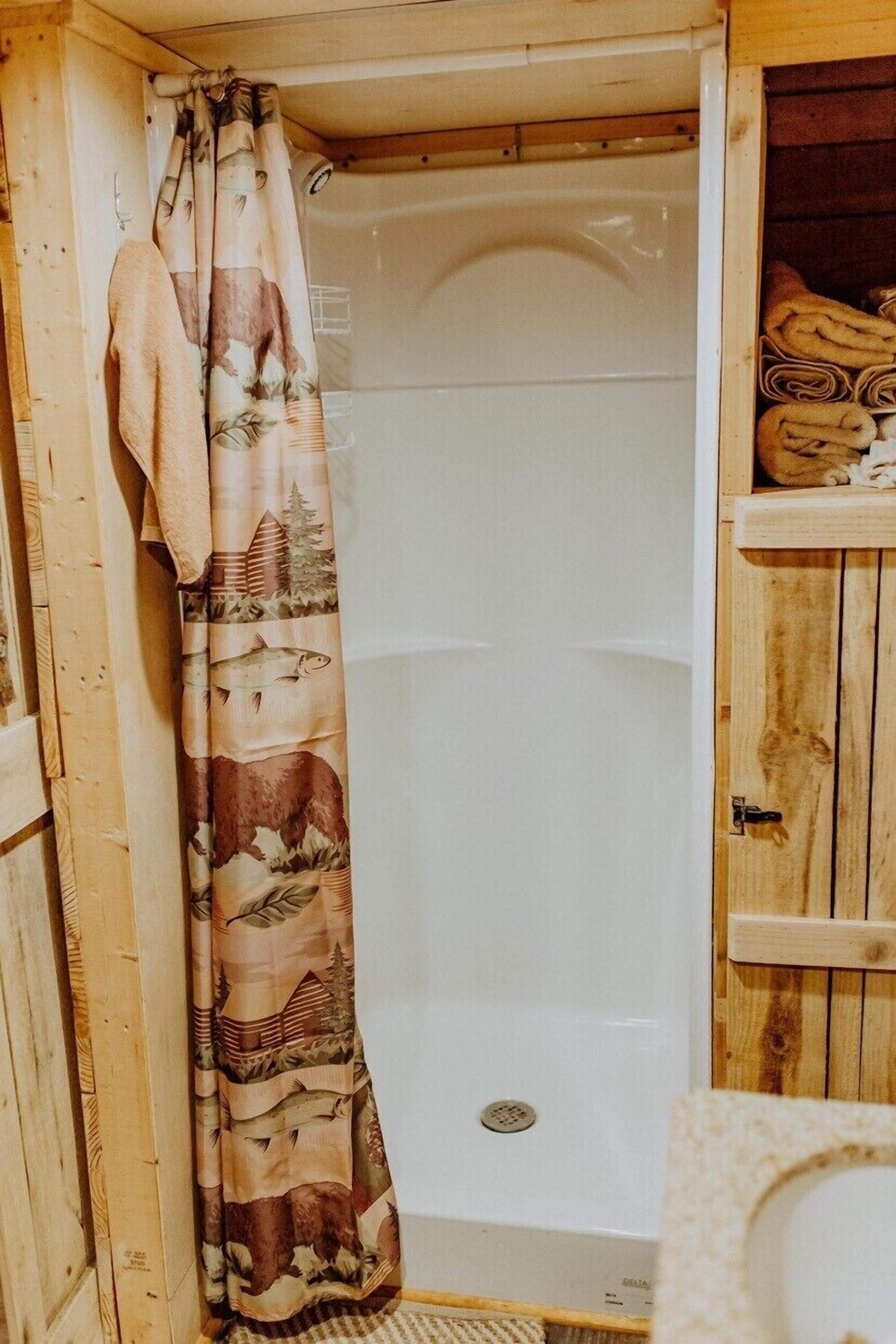 A wooden cabin bathroom with a shower curtain featuring a nature print, a towel hanging on the wall, and a wooden shelf with towels.