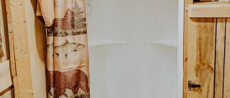 A wooden cabin bathroom with a shower curtain featuring a nature print, a towel hanging on the wall, and a wooden shelf with towels.