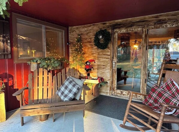A cozy outdoor seating area with a wooden bench and a rocking chair, a Christmas tree, and a wreath on the wall.