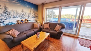 Uma sala de estar acolhedora com um sofá seccional marrom grande, duas almofadas decorativas estampadas, uma mesa de centro de madeira e um mural com tema de inverno na parede.