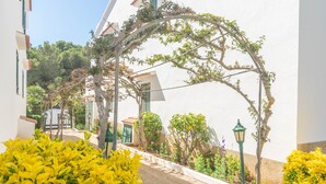 A narrow street with a white building, a green lamp post, and yellow flowering bushes. - SES FONTS C25 - Charming flat with shared pool and close to the beach. Free WiFi. (Es Mercadal)