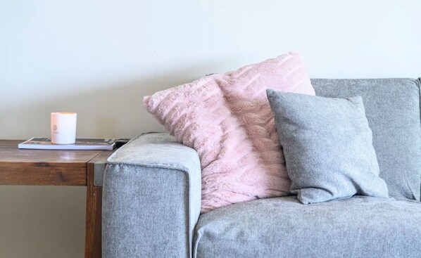 Un sofá gris con un cojín rosa y gris, una vela y un libro sobre una mesita de madera.
