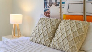 Un lit avec une couverture blanche et deux coussins à texture, une lampe de chevet et une affiche accrochée au mur.