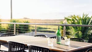 Une table en bois avec des chaises, une bouteille de S.PELLEO et des verres garnis de tranches de citron, sur un balcon offrant une vue sur la verdure et un paysage lointain.