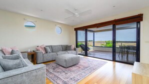 Un salon spacieux avec un canapé gris, une table basse et un grand tapis. Une porte-fenêtre coulissante donne accès à un balcon avec vue sur la plage.
