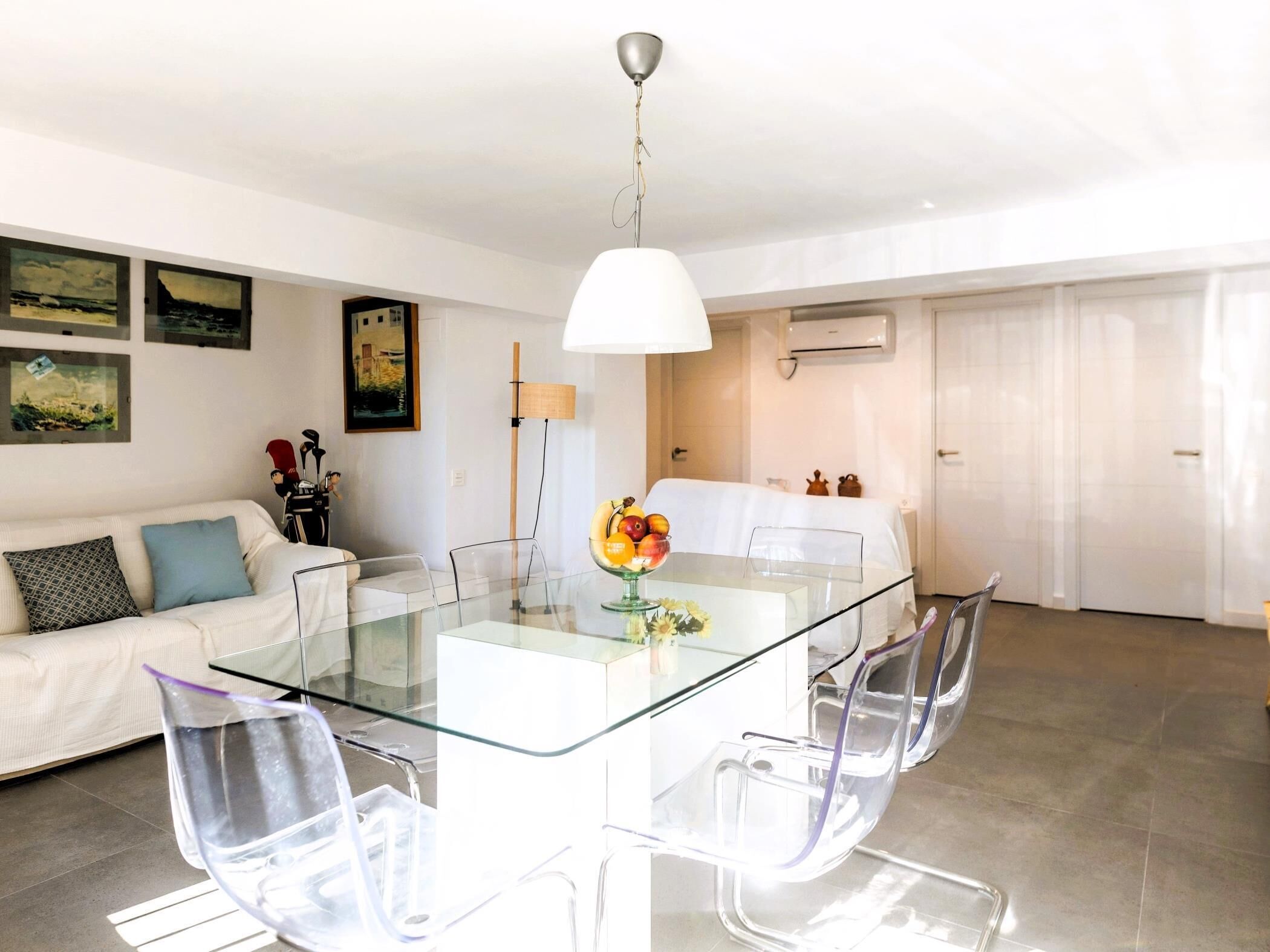 A modern dining area with a glass table and clear chairs, a white sofa with patterned cushions, and artwork on the walls.