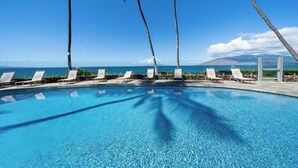 Ein Pool mit klarem, blauem Wasser, umgeben von Liegestühlen und Palmen, mit Blick auf das Meer und die Berge im Hintergrund.