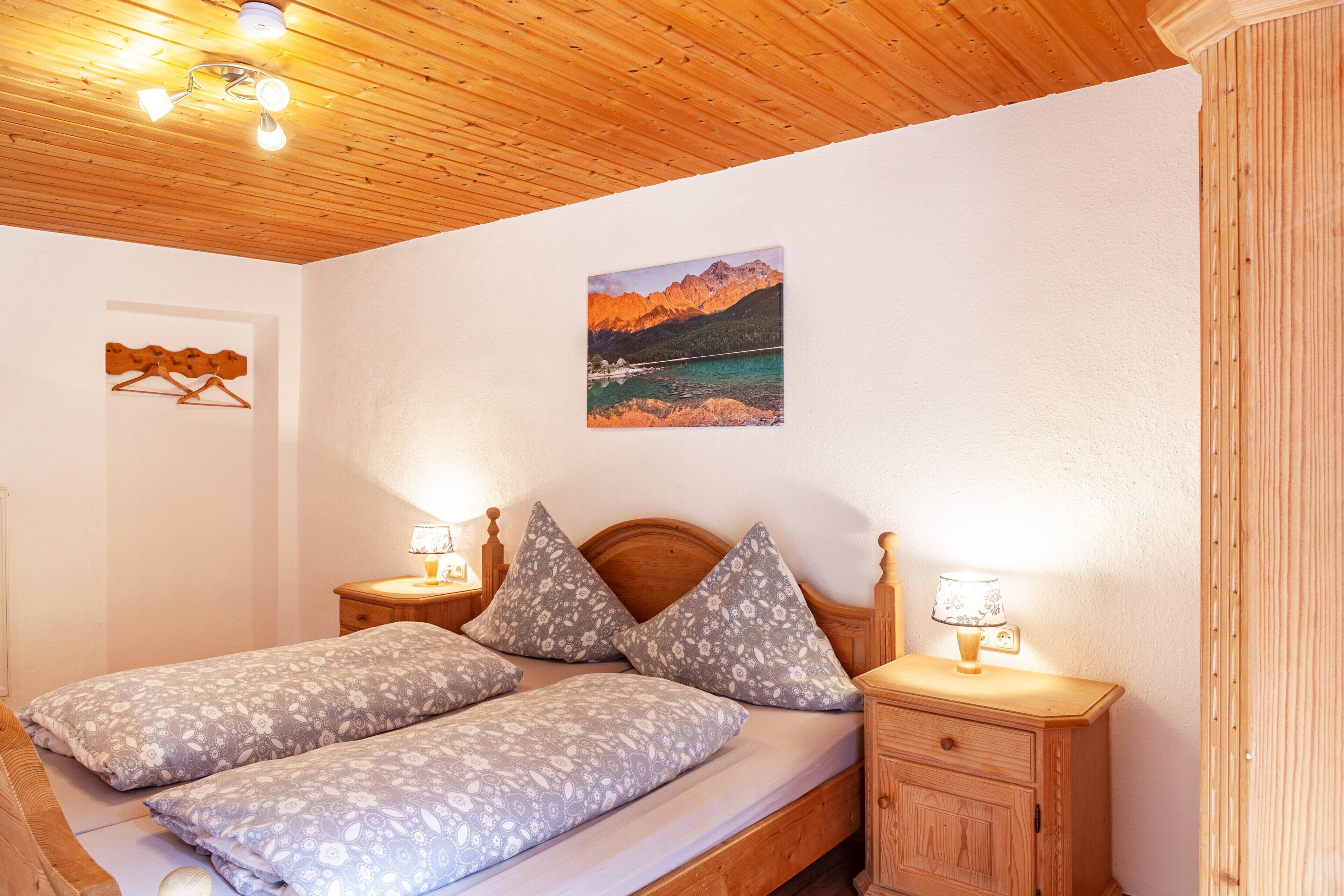 Un plafond en bois, un lit avec une literie  à motifs floraux, une table de chevet avec une lampe et un tableau représentant un paysage de montagne accroché au mur.