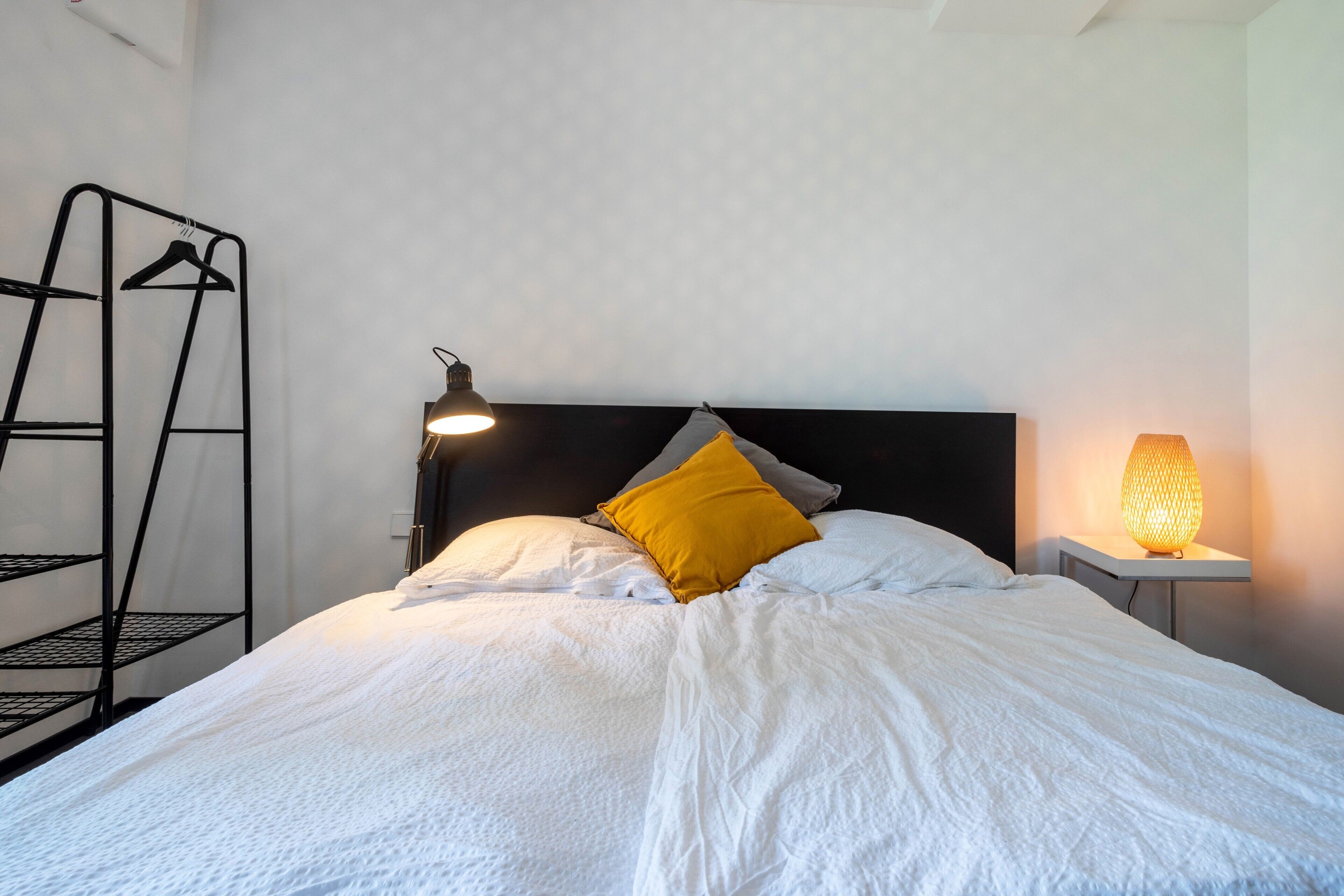 A bed with white bedding, a black headboard, a yellow pillow, a bedside lamp, and a black metal shelf unit.