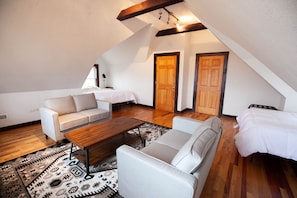 Un dormitorio estilo loft con techo inclinado, una viga de madera en el techo, dos puertas de madera, una cama blanca, un sofá blanco, una mesa de centro de madera y una alfombra estampada.
