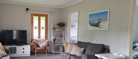Un salon avec un canapé gris, une table d’appoint blanche, un téléviseur, un fauteuil en osier, une bibliothèque et un tableau représentant une scène de plage.