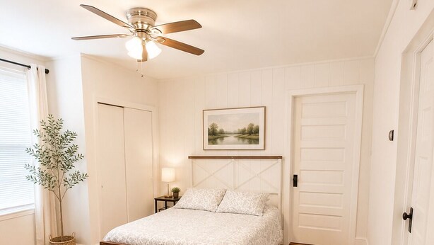 Un dormitorio con cama, ventilador de techo, mesita de noche, lámpara, un cuadro enmarcado en la pared y una ventana con cortinas.