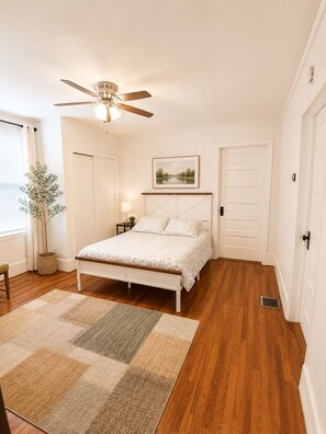 A bedroom with a bed, a ceiling fan, a nightstand, a lamp, a framed picture on the wall, and a window with curtains. - Spacious Thomaston Retreat~Sleeps 10~Families • Work Crews • Weddings (Thomaston)