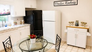 Una cocina con una mesa de comedor de vidrio, sillas, un refrigerador negro, un refrigerador blanco y un armario con un letrero que dice 'Practice Kindness'.