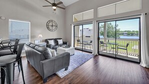 Ein geräumiges Wohnzimmer mit grauem Sofa, einem Couchtisch und einem Essbereich mit Stühlen. Ein großes Fenster bietet Blick auf einen See, und ein Balkon ist mit Gartenmöbeln ausgestattet.