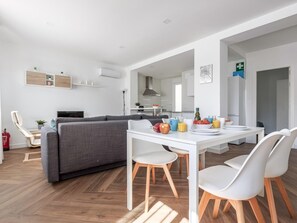 A modern living room with a grey sofa, a dining table with chairs, and a kitchen area in the background.