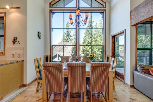 A dining area with a large window, wooden chairs, a chandelier, and a view of trees. - Northern Lights 21 - Ski in Walk out, Private Hot Tub, Fireplace (Whistler)