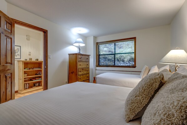 Quarto com cama, penteadeira, mesa de cabeceira, janela com vista para árvores e luminária.