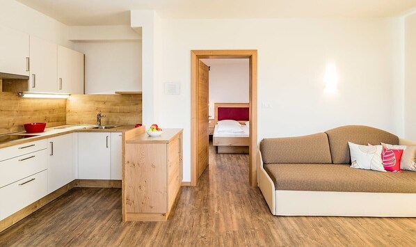 Une cuisine moderne avec des armoires blanches, un comptoir en bois et un saladier rouge. On y voit un tabouret en bois sur lequel est posé un saladier rempli de fruits. Un canapé marron agrémenté de coussins décoratifs se trouve dans l’espace salon. On aperçoit, au fond, un lit avec une tête de lit rouge.