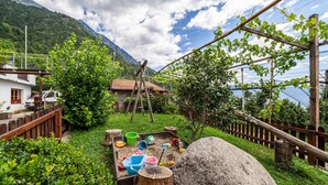 A garden with a wooden play area, a wooden fence, and a house with a red roof. - Holiday Apartment Rasnerhof - Tschigat with Mountain View, Balcony & Wi-Fi (Algund)
