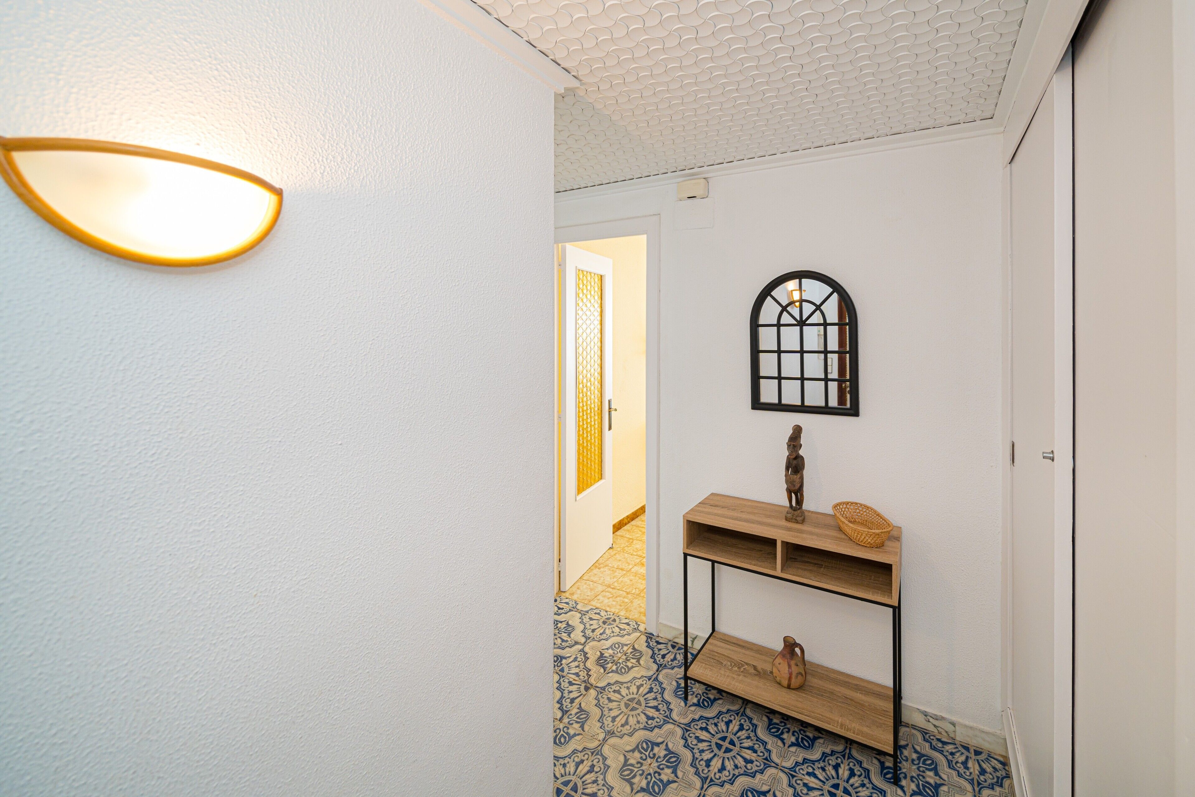 A hallway with a patterned floor, a wall-mounted light fixture, a wooden console table with a basket, and a decorative item.