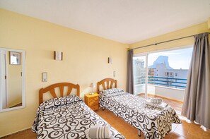 A hotel room with two beds, a small bedside table, a window with curtains, and a view of buildings and a mountain. - Apto en Primera Línea Arenal Calpe with Sea View, Terrace & Wi-Fi (Calpe)