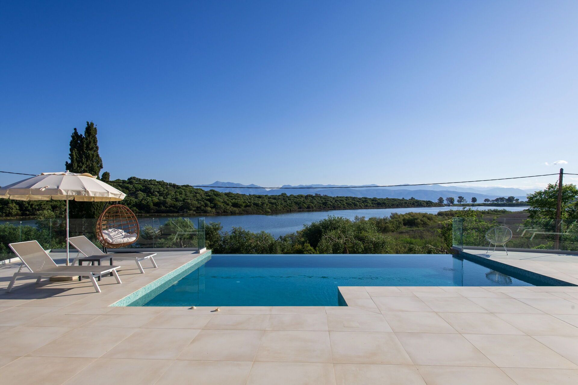 Un espace piscine doté de chaises longues et d’un fauteuil en osier, offrant une vue sur un lac et des montagnes au loin.