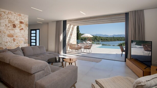 A modern living room with a grey sofa, a TV on a wooden stand, and a view of a pool and mountains through large glass doors.