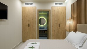 A bedroom with a bed, two wooden wardrobes, a mirror with a circular light, a bedside lamp, and a television on the wall.