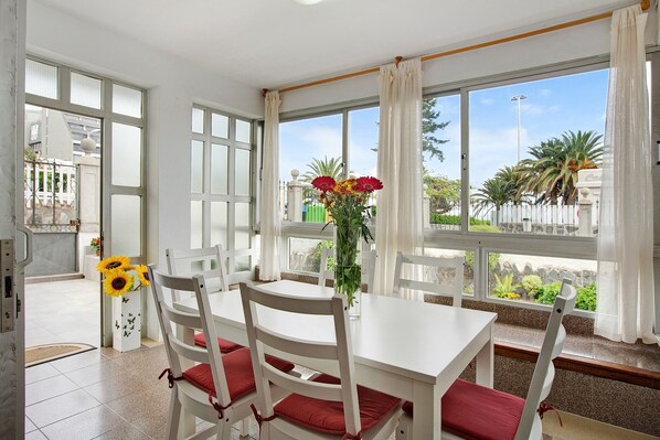 A dining area with white furniture, large windows, and a view of greenery outside.