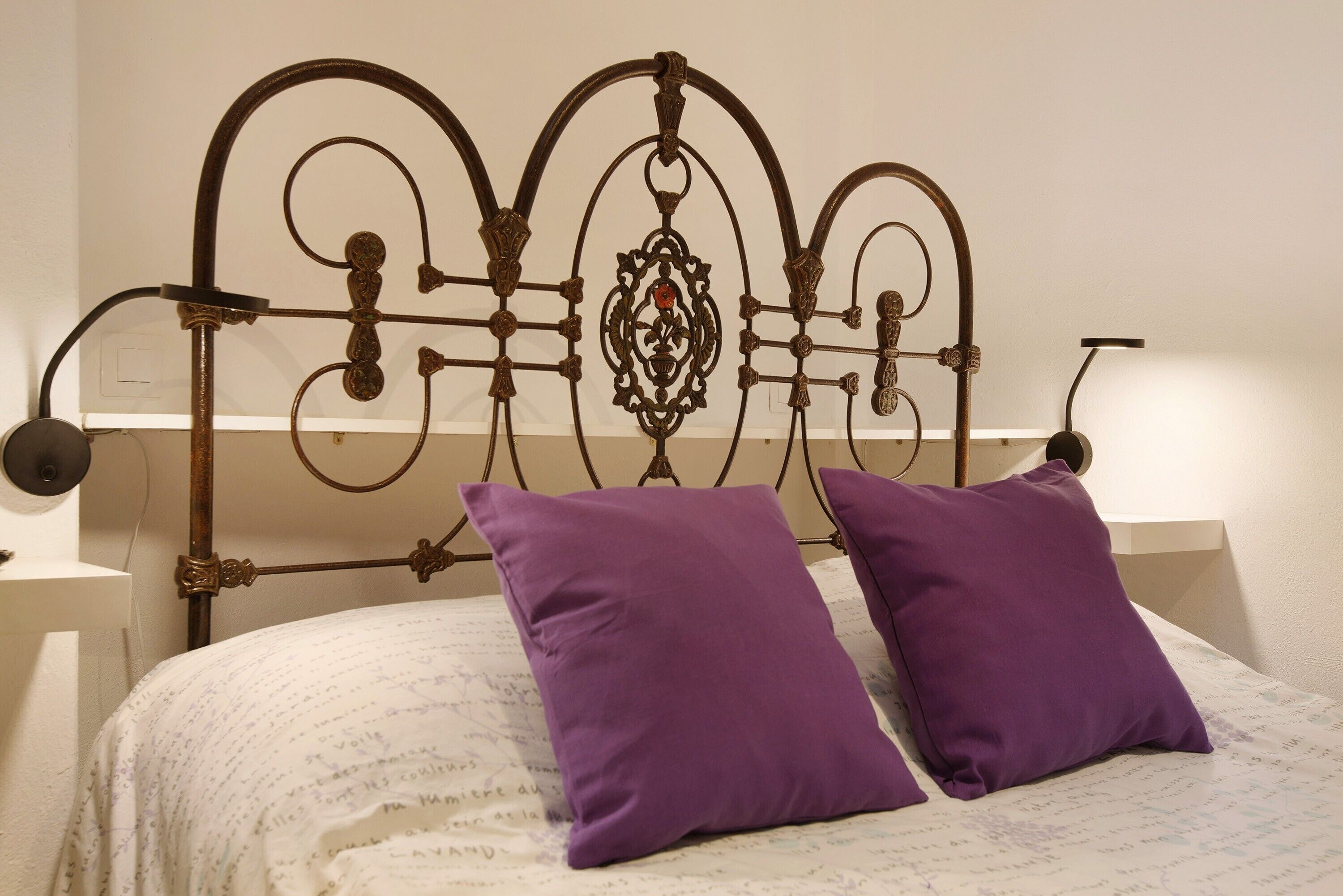 Una cama con cabecera de metal, dos almohadas moradas y una colcha blanca con texto.