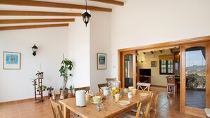 Un coin repas avec une table en bois, des chaises, des plantes en pot et une vue sur l’extérieur grâce à une porte ouverte.